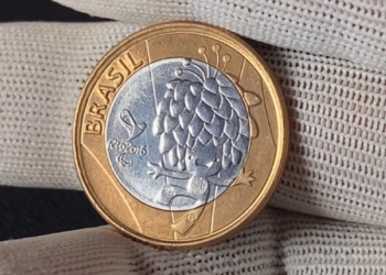 Mão com luva branca segurando uma moeda de 1 Real das Paralimpíadas Rio 2016, apresentando o desenho do mascote Tom em prata sobre o fundo dourado.