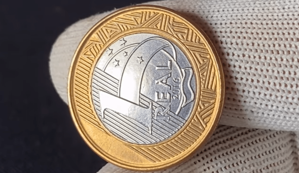 Close frontal de uma moeda de 1 Real das Olimpíadas Rio 2016, focando no mascote Tom prateado e no aro dourado com a inscrição "Brasil".