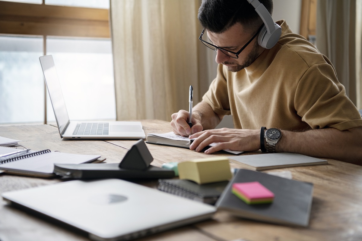 Concurso da Prefeitura Homem jovem usando fones e escrevendo em caderno, laptop aberto e material de estudo sobre mesa de madeira.