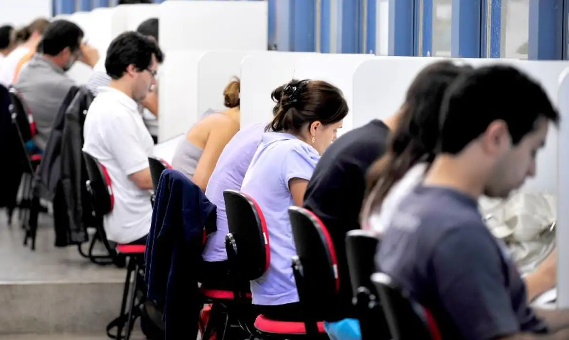 Pessoas sentadas em fileira estudando ou fazendo prova em cabines individuais com cadeiras pretas e vermelhas