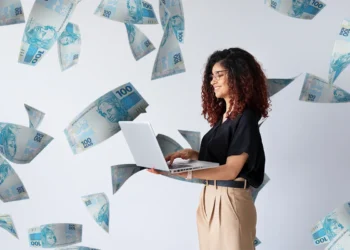 Mulher sorrindo enquanto usa um laptop, com cédulas de 100 reais flutuando ao seu redor, simbolizando o crescimento financeiro em carreiras de sucesso.