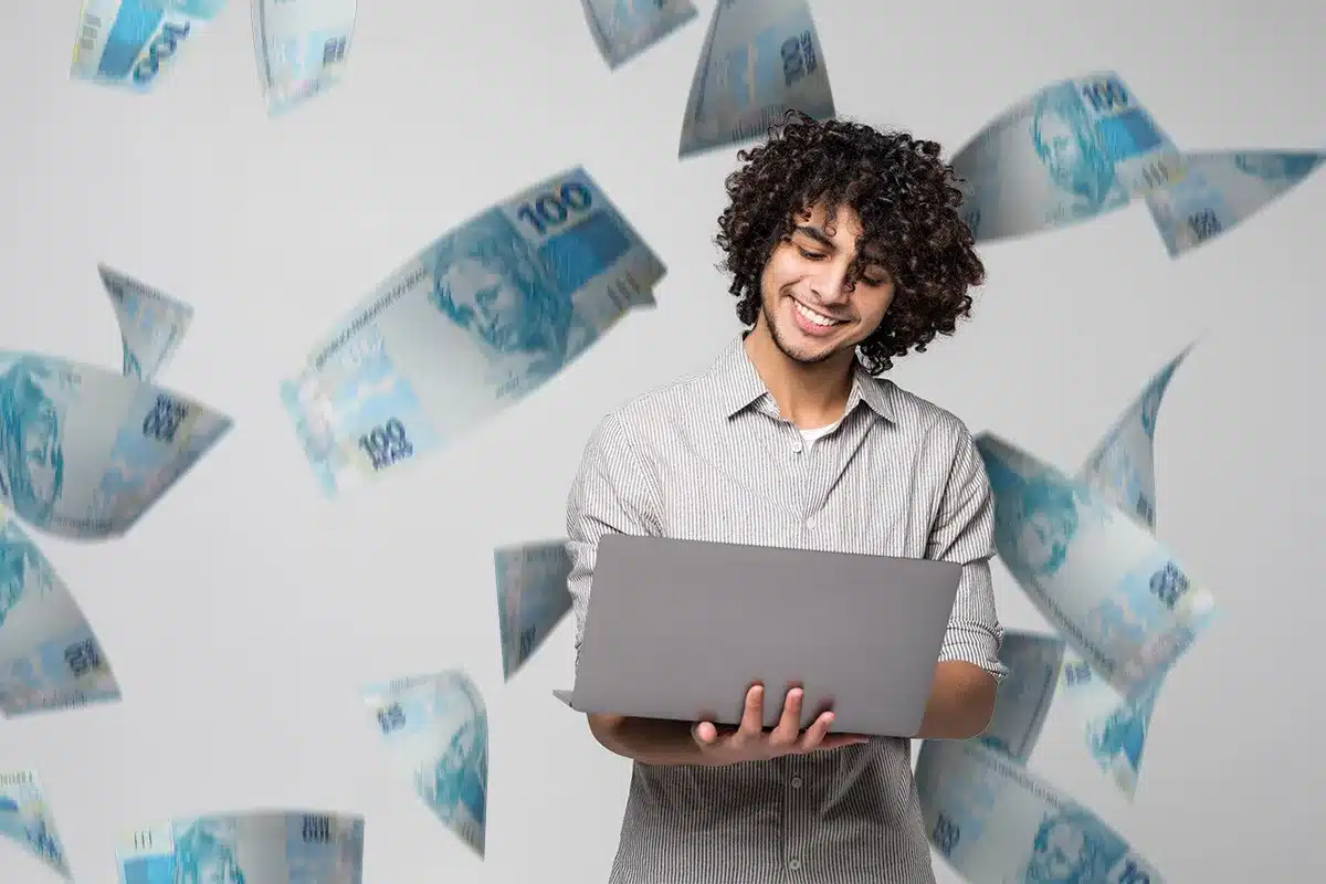 Cargos em alta: Projeções salariais e potencial de crescimento em 2026 Homem sorrindo e usando um laptop, com cédulas de 100 reais flutuando ao fundo, representando o crescimento salarial em cargos em alta.