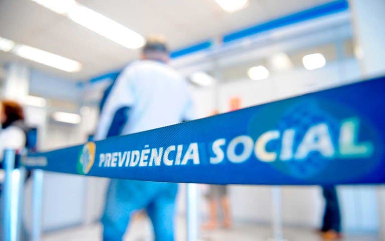 Faixa azul com o logo e o texto "Previdência Social" no balcão de atendimento do INSS, com pessoas aguardando. Representa a mudança nas perícias médicas.