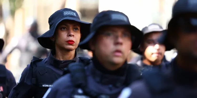 Imagem de policiais militares da Polícia Penal de Minas Gerais em treinamento, com foco em uma mulher com maquiagem e expressão séria, usando uniforme preto e boné característico da corporação.