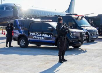 Agentes da Polícia Penal Federal em operação com viaturas e aeronave ao fundo.