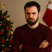 Homem de suéter vinho com expressão mal-humorada, árvore de Natal decorada e meia de Natal vermelha na parede