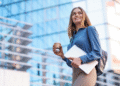 Mulher sorridente segurando laptop e copo de café na mão, com prédios de vidro ao fundo