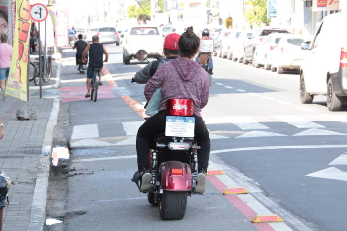 Homem com boné vermelho e mulher com moletom andando de moto elétrica na ciclovia de rua movimentada
