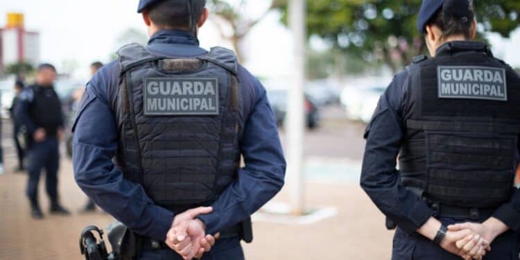 Dois guardas municipais vestidos com uniformes azuis e coletes à prova de balas em postura de vigilância na rua.