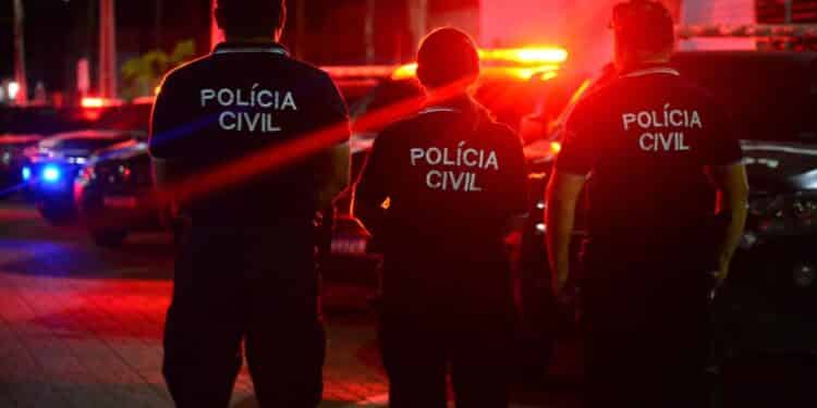 Três agentes da Polícia Civil de costas com viaturas policiais e luzes vermelhas ao fundo à noite