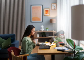 Pessoa concentrada, com uma xícara na mão, estudando em seu escritório, com laptop e materiais de estudo na mesa, ideal para ilustrar a preparação para os concursos de 2026.