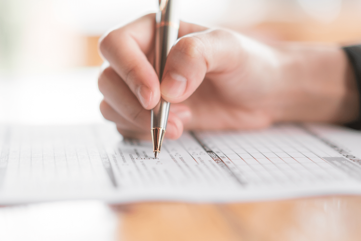 Mão segurando caneta prateada preenchendo dados em formulário impresso sobre mesa de madeira