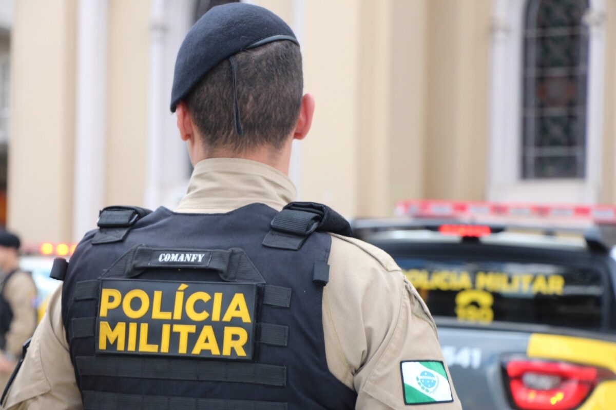 Soldado da Polícia Militar de costas usando colete preto e boné azul, com viatura ao fundo.