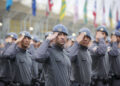 Grupo de policiais militares em uniforme cinza e boina azul realizando saudação em formação