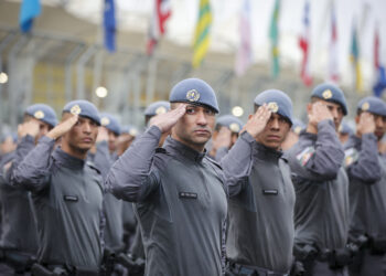 Grupo de policiais militares em uniforme cinza e boina azul realizando saudação em formação