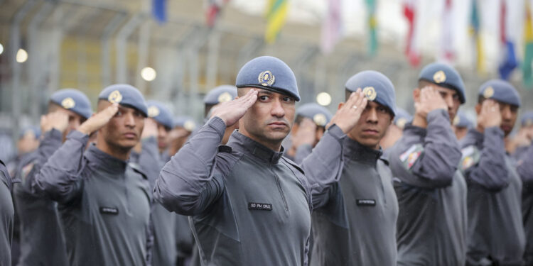 Grupo de policiais militares em uniforme cinza e boina azul realizando saudação em formação