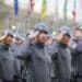 Grupo de policiais militares em uniforme cinza e boina azul realizando saudação em formação