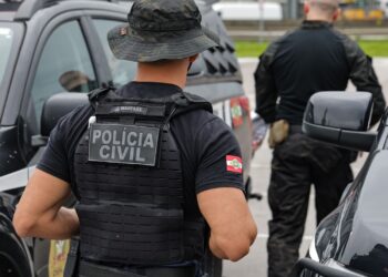 Agentes da Polícia Civil com colete tático preto diante de viaturas durante ação policial.