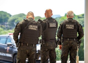 Três policiais penais com uniformes táticos e coletes à prova de balas vistos de costas