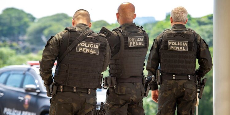 Três policiais penais com uniformes táticos e coletes à prova de balas vistos de costas