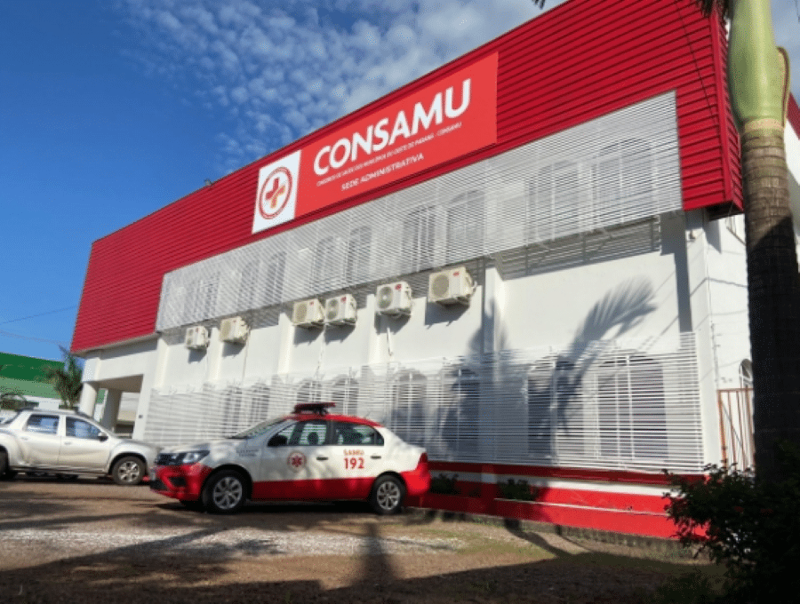 Fachada da sede administrativa do CONSAMU com ambulância do SAMU 192 estacionada em frente, sob céu azul.