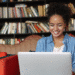 Jovem mulher sorridente usando laptop sentada em sofá vermelho com prateleira de livros ao fundo