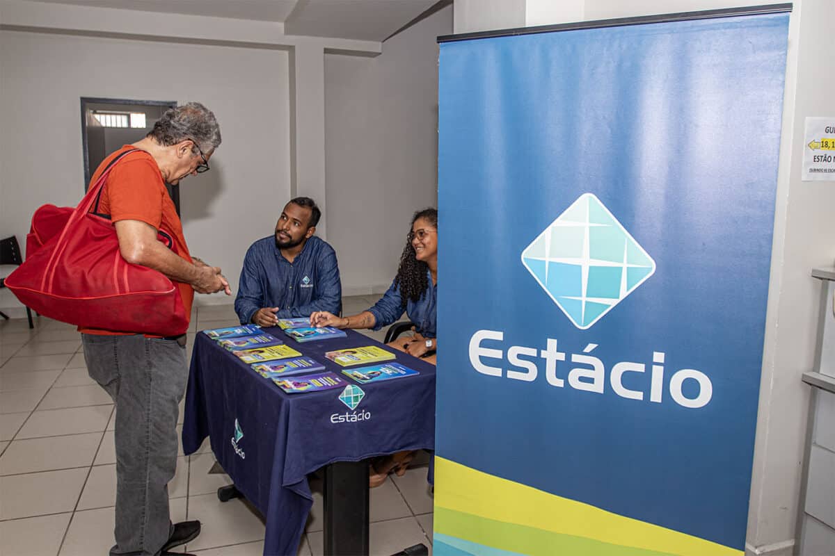 Homem com bolsa vermelha conversa com atendentes da Estácio em mesa com folhetos informativos