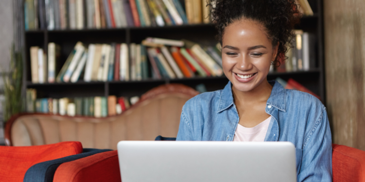 Jovem sorridente usando notebook sentada em poltrona vermelha com estante de livros ao fundo