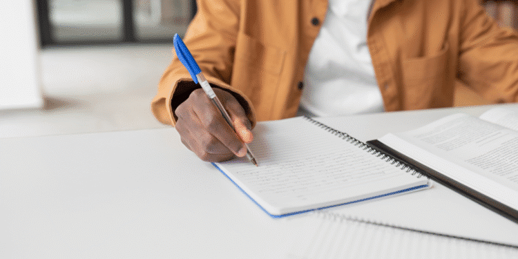 Pessoa escrevendo em caderno espiral ao lado de livro aberto em mesa branca
