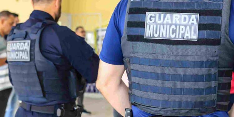 Guardas municipais de costas usando coletes à prova de bala em ambiente interno com outras pessoas ao fundo