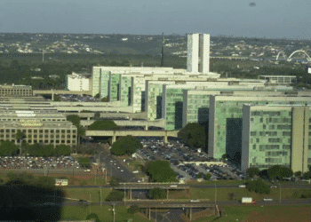 Vista aérea dos prédios do Congresso Nacional com carros no estacionamento e o céu claro ao fundo