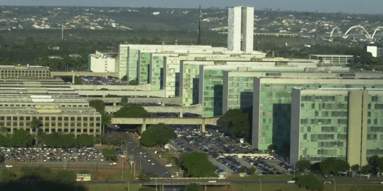 Vista aérea dos prédios do Congresso Nacional com carros no estacionamento e o céu claro ao fundo