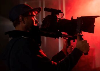 Cinegrafista com boné, filmando com câmera profissional em um set de gravação, iluminado por luz vermelha.