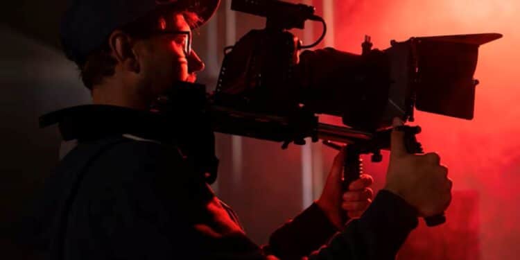 Cinegrafista com boné, filmando com câmera profissional em um set de gravação, iluminado por luz vermelha.