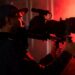 Cinegrafista com boné, filmando com câmera profissional em um set de gravação, iluminado por luz vermelha.