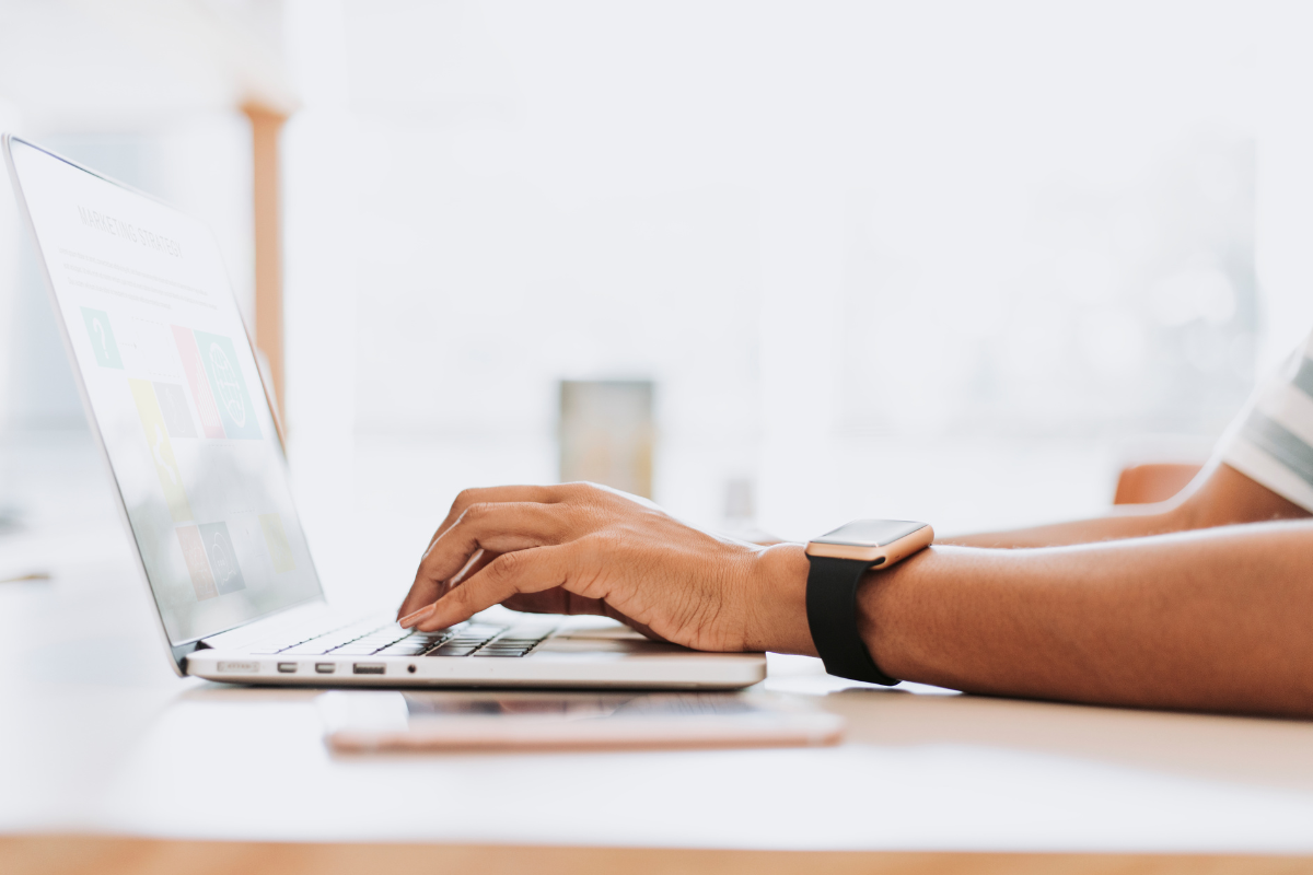 Mão de pessoa com smartwatch preto digitando em notebook prateado sobre mesa branca