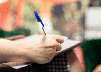 Mão de pessoa escrevendo com caneta azul em caderno apoiado no joelho em ambiente com fundo desfocado