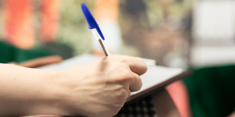 Mão de pessoa escrevendo com caneta azul em caderno apoiado no joelho em ambiente com fundo desfocado