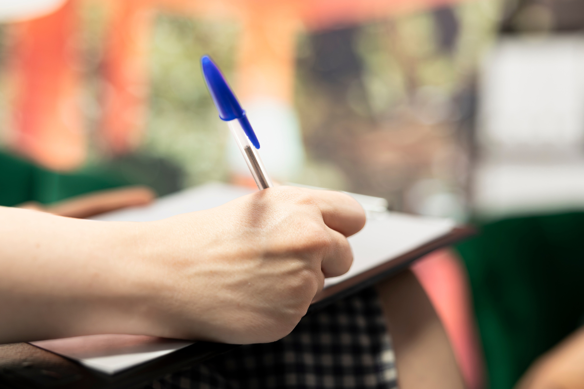 Concursos Prefeituras Mão segurando caneta azul escrevendo em bloco de notas em ambiente desfocado ao fundo