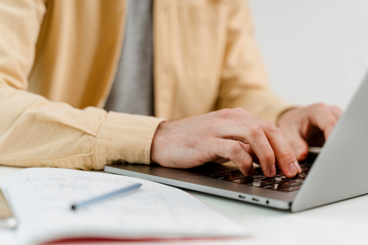 Inscrição vagas de emprego Mãos de pessoa digitando em teclado de laptop prata com camisa bege e caderno aberto com caneta ao lado