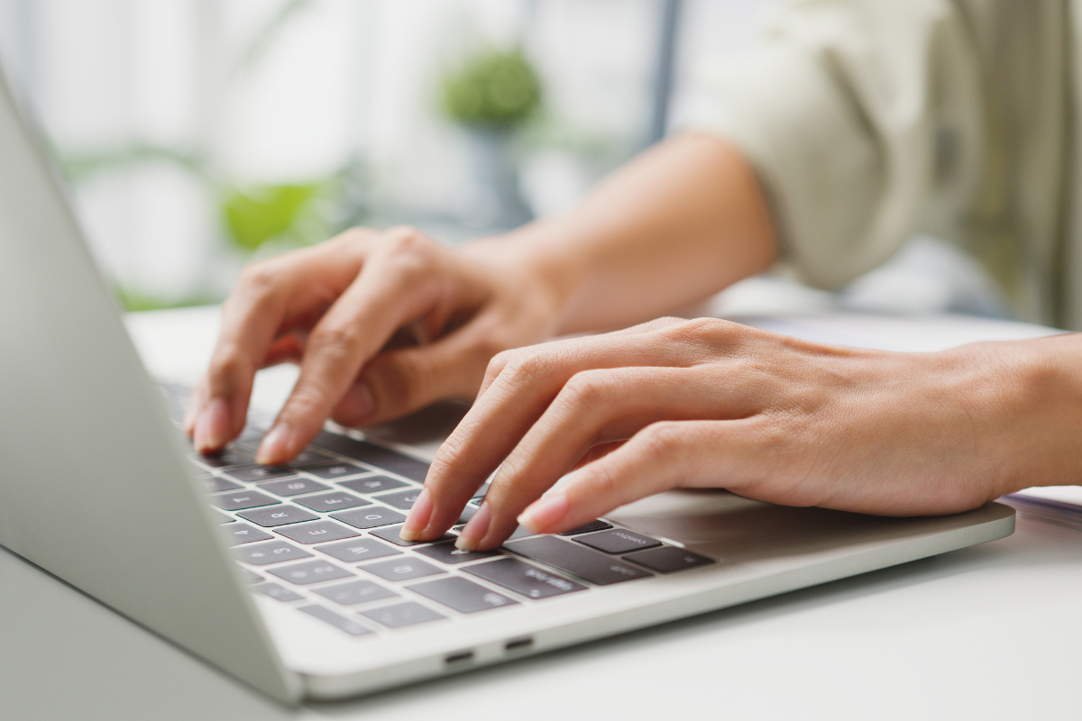Mãos femininas digitando em teclado de notebook cinza em mesa branca com fundo desfocado