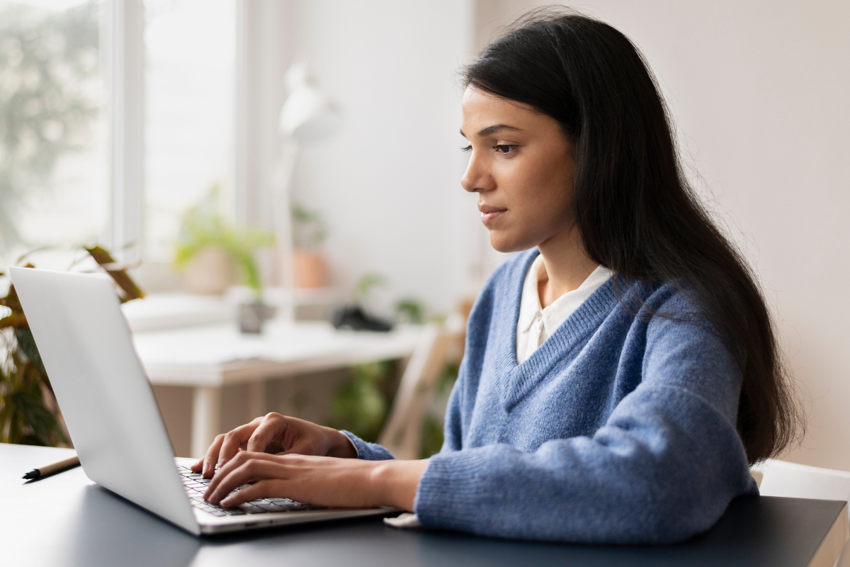 Mulher usando suéter azul, digitando em laptop cinza em mesa com fundo desfocado e luz natural