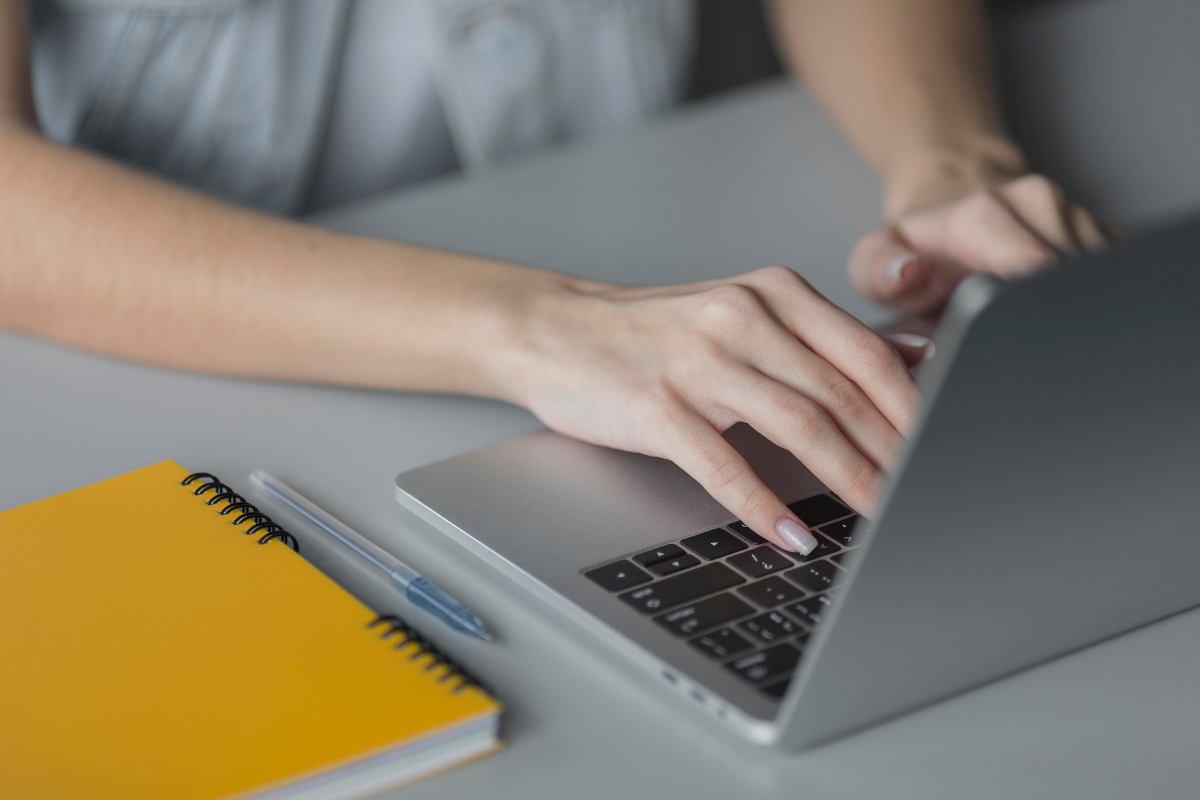 Mãos femininas digitando em notebook cinza próximo a caderno amarelo e caneta azul sobre mesa cinza