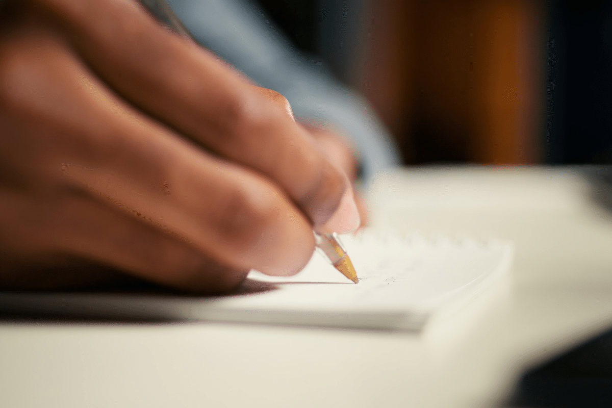 Imagem de uma mão segurando uma caneta esferográfica amarela e escrevendo em caderno branco em superfície clara.