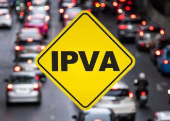 Placa amarela de trânsito em formato de losango com a sigla IPVA escrita em preto, sobreposta a uma imagem desfocada de congestionamento de carros à noite com luzes vermelhas acesas.