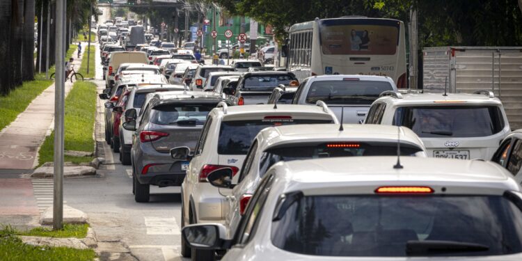 Trânsito congestionado com carros parados em fila dupla em via urbana arborizada durante dia claro
