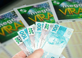 Mão segurando cédulas de dinheiro com bilhetes da Mega Virada ao fundo.