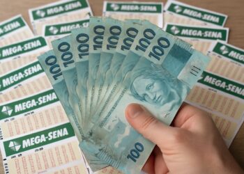 Pessoa segurando notas de cem reais com bilhetes da Mega-Sena ao fundo