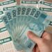 Pessoa segurando notas de cem reais com bilhetes da Mega-Sena ao fundo