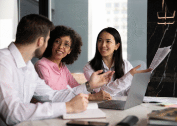 Grupo de três profissionais discutindo documentos e ideias em ambiente corporativo com laptop aberto